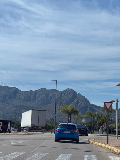 The image shows a street in Ondara, Spain, with a mountainous backdrop and a clear sky with scattered clouds. A blue car with license plate "1222 LRC" is parked on the right side of the road, followed by a gray vehicle. A white truck is further down the road. Traffic signs are visible along the roadside. The road has zebra crossings. The landscape includes palm trees and other greenery. The word "ONRA" can be seen on a sign.