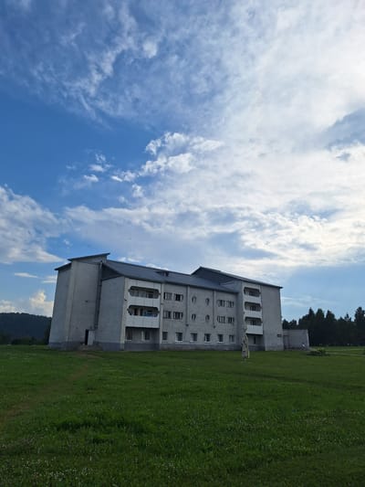 A wide-angle, low-angle shot captures a long, white, three-story building situated in Atamanovka, Russia. The building has a dark, sloped roof and features balconies with white railings. The facade of the building is punctuated by numerous windows, some of which are circular and others rectangular. In front of the building, a vast expanse of green grass, dotted with small white flowers, stretches towards the viewer. In the distance, a line of dark green trees forms a backdrop, and a gentle, forested slope rises behind them. The sky above is a vibrant blue, adorned with scattered white clouds, some of which are thin and wispy, while others are more substantial and fluffy. The overall lighting suggests it is daytime, likely late afternoon or early evening, with the sun positioned towards the upper right of the frame. A solitary figure, appearing to be a person, stands near the building amidst the grass, facing away from the camera. There is also a tall, abstract sculpture made of fabric or similar material positioned in the grass between the building and the viewer.