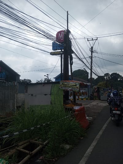 A street scene shows a cluttered roadside. Overgrown grass and debris fill the foreground. A makeshift fruit stand is visible. Overhead, a tangled web of power lines obscures a cloudy sky. Signs for "Gaco" and "Mie Pedas No." adorn a utility pole. A motorcycle with two riders passes on the asphalt road to the right. Other shops and buildings line the street further down the road.