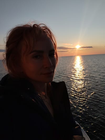A medium shot photograph captures a woman with reddish-blonde hair looking towards the camera. She is positioned on the left side of the frame, with her face partially illuminated by the setting sun. Behind her, a vast body of water stretches to the horizon, reflecting the warm golden hues of the sunset. The sun itself is a bright, central point of light in the upper right quadrant of the image. Wispy clouds are visible in the sky, tinged with orange and pink. The woman is wearing a dark jacket. The scene appears to be an outdoor setting, possibly on a boat or a pier, overlooking the sea at dusk. The location is specified as Kakkarovo, Russia.