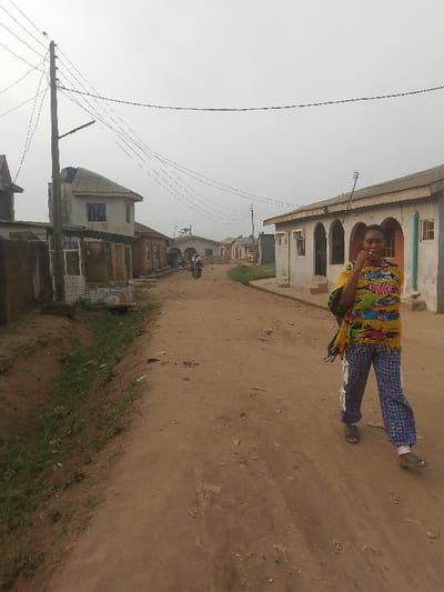 A dirt road lined with simple, low-rise buildings dominates the scene under an overcast sky. Utility poles and wires stretch overhead. A woman in a colorful shirt and patterned pants walks towards the viewer on the right side of the road. In the distance, a person on a motorbike moves away. Vegetation grows along the side of the road. The buildings have arched windows and simple architectural details. The overall impression is of a rural or underdeveloped area.