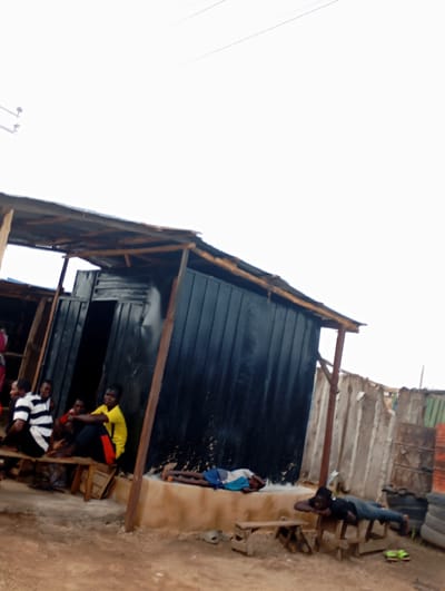 In Shendam, Nigeria, a low-angle, vertical shot captures a scene outside a weathered, corrugated metal structure with a makeshift roof of corrugated tin and wood. In the foreground, a young person lies prone on a wooden bench, head resting on their arms. To the left, a group of four young people sit on another wooden bench. One person wears a black and white striped shirt, another a red shirt, and a third a bright yellow t-shirt with red accents on black pants. A fourth person is partially obscured. The ground is dirt and uneven. Further back, a rough wooden fence and stacked tires are visible. The sky is overcast and bright.