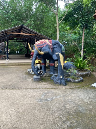 The image shows a large elephant statue made of concrete, standing on a paved area in a small garden surrounded by trees and vegetation. The elephant's trunk is raised, its body is painted black with multi-colored decorations, and its legs are painted in a combination of yellow, red, and white. The elephant is positioned near a large tree trunk, also made of concrete and painted in a dark color. The ground is made of concrete, and there are a few scattered rocks and potted plants. The area is located in a small park or garden, surrounded by trees and vegetation. The location is in Kecamatan Manyaran, Indonesia (latitude: -7.826, longitude: 110.816).