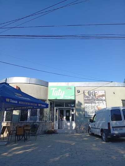 A bright, sunny day in Buțeni, Moldova, is captured in this eye-level, exterior shot of a market with outdoor seating. The building's facade is painted a muted green, with a curved silver-colored section on the left. Above the entrance, a green sign reads "Taty market" in white lettering, with a smaller "TYMBARK" logo above it. To the right of the entrance, a large banner displays an image of beer taps with the text "BERE CRAFT" and "TATY MARKET" below.
In front of the building, a blue umbrella from "Chisinau Bere Mereu Intre Prieteni" provides shade over a set of wooden tables and chairs. The ground is paved with cobblestones, showing some cracks. A white Opel van with the license plate "MD JUV 650" is parked to the right of the entrance. The sky is a clear, vibrant blue, with several dark power lines crossing the frame from left to right. The overall scene suggests a commercial establishment in a semi-urban or rural setting.