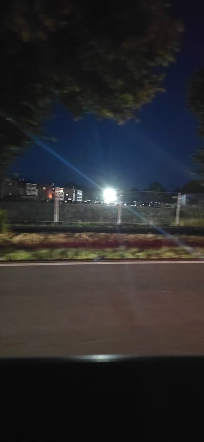 The image shows a view from a moving vehicle at night, likely on a road adjacent to a residential area. In the background, several multi-story buildings are visible, illuminated by artificial lights. A bright light source, possibly streetlights or a powerful car headlight, is intensely shining, creating lens flare and beams across the dark blue night sky. Trees with foliage are partially obscuring the top of the frame, suggesting an outdoor setting in a semi-urban or suburban environment. A metal fence runs horizontally across the mid-ground, separating the road from the buildings and a strip of vegetation. The foreground consists of a dark asphalt road with a white dividing line. The overall impression is that of a nighttime journey through a town or city. The location of Neustadt-Glewe, Germany, is not explicitly depicted or identifiable within the image itself, as there are no specific landmarks or signs that would indicate this particular place.