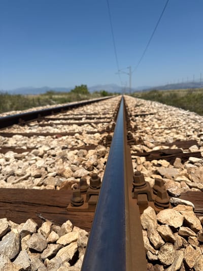 The image is a low-angle, close-up shot of a railway track stretching into the distance. The perspective is from beside the right-hand rail, focusing on the metallic sheen of the track itself and the surrounding ballast, which consists of rough, light-colored stones. Wooden railway ties are visible beneath the ballast, some appearing weathered. Metal fasteners, including bolts and nuts, are securing the rail to the ties.

In the background, the railway line diverges into two sets of tracks, each lined with similar ballast. Overhead, power lines for an electric railway are suspended from poles, creating thin lines against a clear, bright blue sky. Beyond the tracks, the landscape consists of gently rolling hills covered in dry, green and brown vegetation. Distant mountains are faintly visible on the horizon. The scene suggests a rural or semi-rural setting, possibly a railway line in an open area. The location is specified as Mojanovići, Montenegro. There are no people or animals visible in the image. The overall impression is one of a clear, sunny day.