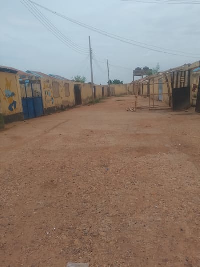 The image depicts a view of a dirt road in front of a row of low-lying buildings, likely a school, in Gindi Nassarawa Kyarmi Galbi, Nigeria. The buildings are painted a light yellow and have metal roofs. One building on the left has murals of cartoon characters painted on its wall, including a blue character with large ears. A blue gate is visible on this building. Further down the row, there are more doors and some unpainted sections of wall. A metal gate with vertical bars is seen in the middle right of the image. Above the buildings, utility poles and wires are visible against an overcast sky. In the background, some trees and water tanks on a raised platform can be seen. The foreground is dominated by the unpaved, sandy road with scattered pebbles and debris. There are no people or animals visible in the image.