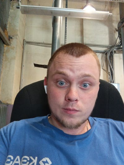 The image shows a close-up, selfie-style shot of a fair-skinned man with light brown hair and blue eyes, wearing a blue t-shirt and a silver chain necklace. He has a small goatee and is wearing white wireless earbuds. He is seated in what appears to be a dark chair, with a concrete and metal industrial-looking environment in the background, featuring exposed pipes, electrical conduits, and lighting fixtures. The specific location within the city of Kursk, Russia, cannot be determined from the visual information present.
