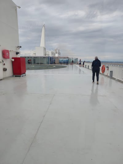 The image depicts the open deck of a ferry with a light-colored surface, likely the bow or stern, extending into the foreground. A person with gray hair, wearing a dark jacket and pants, is walking away from the camera towards the right side of the frame. In the middle ground, several other individuals are visible, some standing and others appearing to observe something on the deck. A red fire extinguisher cabinet is mounted on the left side of the white structure that forms the edge of the deck. Further back, a helipad marked with yellow symbols is visible on a green surface, adjacent to enclosed passenger areas with large windows. The deck is wet, reflecting the overcast sky. In the background, a railing separates the deck from the sea, with land visible on the horizon. The environment suggests a maritime setting, possibly Apallund, Åland, given the ferry context and the possibility of this being a common route.