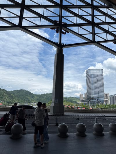 The image depicts a partially obstructed view of Yongding District, Zhangjiajie, China. A modern skyscraper rises in the background under a cloudy sky. Foreground elements include a geometric metal structure, possibly part of a transportation hub's roof, and concrete pillars with spherical tops. A small group of people is visible, seemingly waiting with luggage. The scene suggests a transportation terminal overlooking the city's landscape.