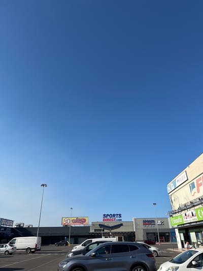 The image depicts a sunny day in the city of Sedaví, Spain, likely a commercial area, with a clear blue sky dominating the upper portion. In the foreground, a parking lot is filled with various vehicles, including white vans, sedans, and SUVs. The buildings in the background feature prominent signage for businesses such as "SPORTS DIRECT.COM," "USC," and "Chiosofas." One building has a large banner that reads "FLEX" and displays promotional text in Spanish. A tall lamppost stands in the middle ground. The overall scene suggests a retail or commercial hub.