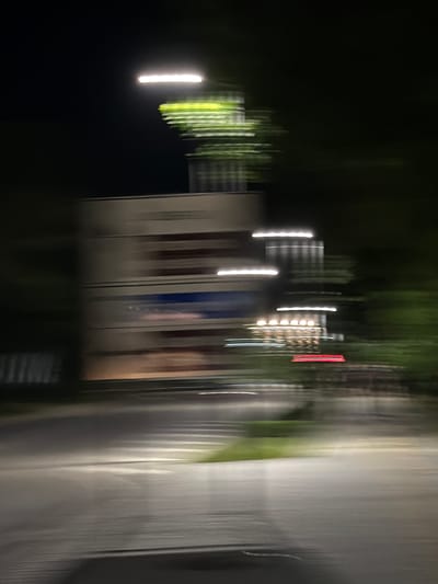 The image captures a motion-blurred nighttime scene in the city of Pravets, Bulgaria. A large, horizontally oriented sign with horizontal stripes, possibly advertising, is visible on the left side of the frame. Above the sign, blurred green foliage and a bright horizontal light suggest street lighting or illumination from the structure. To the right of the sign, a series of horizontal white lights are streaked across the frame, and lower down, a cluster of circular yellow lights and a red rectangular light are also visible, indicative of building lights or signage. The foreground shows a blurred road surface with white lines and patches of green grass, suggesting the image was taken from a moving vehicle. The overall impression is that of a drive-by view at night, with the motion blur obscuring fine details but highlighting the illuminated elements of the urban landscape.