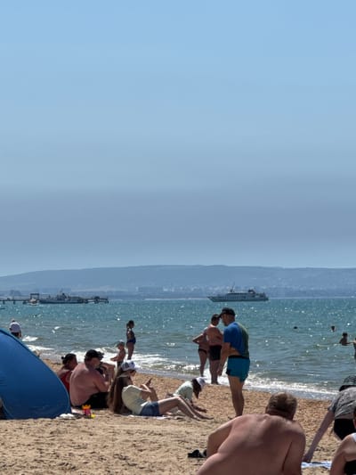 The image depicts a sunny day at a crowded beach in Vityazevo, Russia. The scene is dominated by a sandy shoreline with a clear, blue sky above. The sea, sparkling under the sun, is a prominent feature, with gentle waves lapping at the shore. A large ferry or ship is visible in the distance, sailing on the calm water.

Several groups of people are scattered across the beach, engaged in various activities. Some are sunbathing on towels and blankets, while others are wading in the shallow water. A few individuals appear to be exercising or stretching. The atmosphere suggests a leisurely and enjoyable day at the beach, with a sense of relaxation and recreation.

The people are dressed in swimwear, appropriate for a warm day. The time of day appears to be midday or early afternoon, given the bright sunlight and clear sky. There are no visible signs or text in the image. The overall impression is one of a typical summer day at a popular seaside resort.
