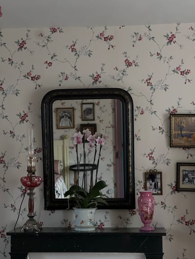An interior room in Claude Monet's house in Giverny, France, is depicted. The walls are covered in a light-colored wallpaper featuring a repeating pattern of intertwining branches with small white and reddish-pink flowers and green leaves. A dark, ornate mirror with a black and gold frame is centrally positioned above a dark, possibly black, mantelpiece.

On the mantelpiece, from left to right: a vintage-style oil lamp with a clear glass chimney, a red glass reservoir patterned with white flowers, and an elaborate silver-colored metal base is situated. A black power cord extends from the lamp's base. In the center, a potted Phalaenopsis orchid with multiple pink blooms and green leaves rests in a white ceramic pot adorned with delicate orange and green bird and floral motifs, sitting on a matching saucer. To the right, a pink, somewhat iridescent vase with a flared rim and base, featuring subtle decorative patterns, completes the display.

The mirror reflects parts of the room opposite, including a light-colored piece of furniture (possibly a bed or chest of drawers) and a partial reflection of an individual or draped object in the lower left quadrant of the mirror. Several framed pictures are also reflected within the mirror: one depicting a man with a white beard and hat, and another showing a landscape or building.

To the right of the mirror, on the wall, a gold-framed painting of yellow flowers in a vase alongside fruit is hung above two smaller, darker-framed photographs. The upper photograph appears to show two individuals in period clothing.