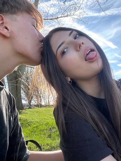 A photograph, oriented on its side, depicts two individuals in an outdoor setting. The person in the upper portion of the frame is a female, and she is facing the camera, her head tilted back, with her tongue visible. She has dark hair, fair skin, and dark eyebrows. The person in the lower portion of the frame appears to be kissing her. They have short hair and fair skin. The background consists of trees and grass, suggesting a park or natural area. The sky is visible in the upper left corner. It is partly cloudy. The image appears to be taken during the day, with natural light illuminating the subjects. The city of Beli Brod, Bulgaria is not discernible.