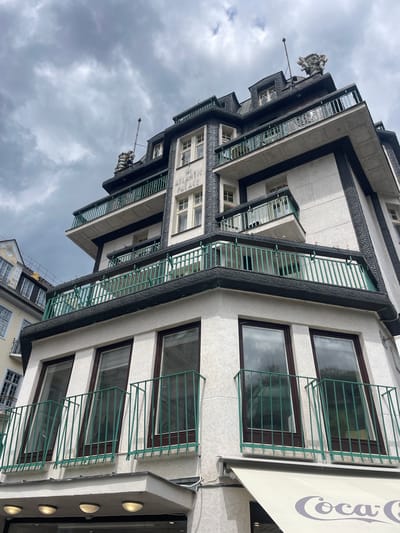 The image depicts the Hotel Atlantic Palace, located in Karlovy Vary, Czechia, under an overcast sky. The multi-story building exhibits a distinctive architectural style, characterized by a light-colored, textured stucco facade contrasted with prominent dark-colored, ornate trim. This trim features a continuous, intricate embossed pattern, framing windows and outlining the structure's angular elements and multiple balconies.

Each level of the building features balconies with light green metal railings, often with small pink flowers in planters. Windows are rectangular with dark frames, some appearing as multi-pane casements on upper levels, while lower windows are taller, single-pane units. The building's upper section includes a mansard-style roof with dormer windows, also trimmed in dark, decorative material.

Crowning the highest point of the roofline is a complex sculptural group, appearing to be multiple intertwined figures, possibly cherubs or mythological beings. A tall, slender flagpole extends vertically from the roof adjacent to this sculpture.

At the street level, a white fabric awning with a partially visible "Coca-Cola" logo extends from the building. Three circular, illuminated light fixtures are recessed into the underside of a small overhang above the awning. A portion of an adjacent, lighter-toned building with different architectural details, including a gilded crest resembling a lion or griffin, is visible on the far left. No people are present in the scene.