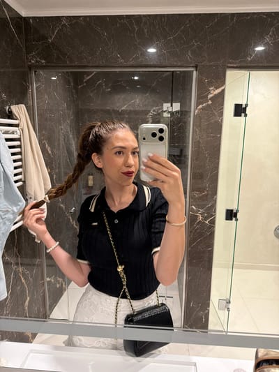 A woman is taking a selfie in a bathroom. She is wearing a black polo shirt with white trim and white lace shorts. Her hair is pulled back in a high ponytail with a braid. She is holding her phone in her right hand and touching her braid with her left hand. A black purse with a gold chain is slung over her shoulder. A white pearl bracelet is on her left wrist. The bathroom has marble walls and a glass shower door. A towel rack with towels is visible on the left. The scene is located in Hisarya, Bulgaria.
