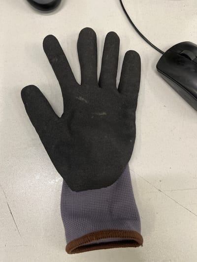 A black glove lies on a textured gray surface. The glove is open-palmed, with fingers extended upward. The cuff of the glove is gray fabric with a brown trim. A black computer mouse and a portion of a black computer tower are visible in the background. The image was taken at an undisclosed location in Novogorelovo, Russia.