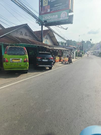 The image shows a street scene in Purwakarta, Indonesia, at the coordinates -6.561 latitude and 107.435 longitude. The scene is shot from the perspective of a vehicle on the road, looking towards the horizon. The road is paved with grey asphalt and has a white lane divider marking the center of the road. On the left side of the road are several commercial buildings, with one building having an awning with a sign that reads "Nasi Isola" in Indonesian. There are two cars parked on the side of the road - a green minivan and a black SUV. A person can be seen in the background riding a motorcycle on the road. The sky is blue and clear with a few white clouds visible. There are also many wires strung across the scene, attached to the buildings and utility poles. The scene is taken from the perspective of a passenger in a vehicle, with the bottom of the vehicle partially visible in the foreground. The image is in focus and well-lit, with a slight sense of motion from the movement of the vehicle from which the photo was taken.