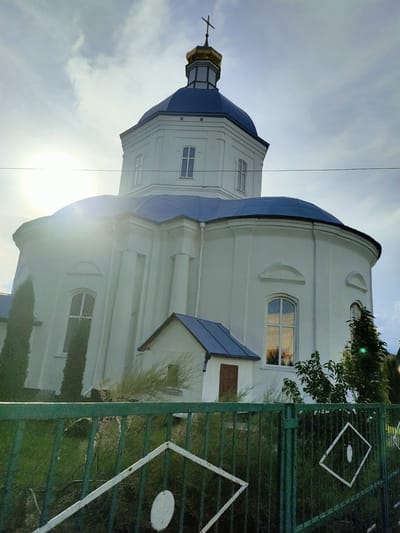 A low-angle, eye-level shot captures the bright white facade of an Orthodox church with a blue domed roof in Nosivka, Ukraine. A golden cross tops the main dome. The sun is strong, creating lens flare to the left of the frame and casting light on the building and the sky, which is partially obscured by clouds. A green metal fence with white diamond and circle decorative elements runs across the foreground. Cypress trees flank the church on the left, and other trees and greenery are visible around the structure, including a small attached building with a blue roof and a brown door. Electrical wires run horizontally across the upper portion of the image. The overall scene depicts a religious building in an outdoor setting.
