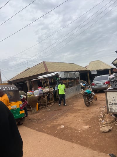This image depicts a street scene in Bokkos, Nigeria, likely in a market area. The foreground shows a portion of a yellow and green auto-rickshaw. Behind it, several market stalls are visible, constructed with corrugated metal and wood, topped with weathered, tan-colored roofs. One stall displays fruits like tomatoes and gourds, along with buckets and a large broom.
A man wearing a bright green t-shirt and black pants stands near a stall, looking towards the viewer. Another individual, partially obscured, is visible near the auto-rickshaw. In the background, a silver sedan is parked, and a motorcycle with bags strapped to its rear is also present. The ground is unpaved, appearing to be dirt and mud. Power lines crisscross the overcast sky, which is filled with gray clouds. The overall impression is one of a bustling, everyday commercial environment in an African town.