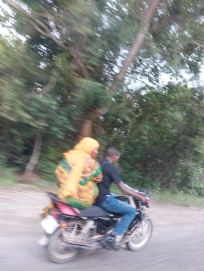In Kisanga, Tanzania, a man and a woman are depicted riding a black motorcycle on a road, captured with significant motion blur suggesting movement. The male driver wears a dark short-sleeved shirt and blue jeans. The female passenger is seated behind him, wearing a yellow head covering and a patterned garment in shades of yellow, brown, and green. A rear license plate is visible on the motorcycle. The background consists of heavily motion-blurred green foliage and tree trunks, indicating a tree-lined roadside environment.