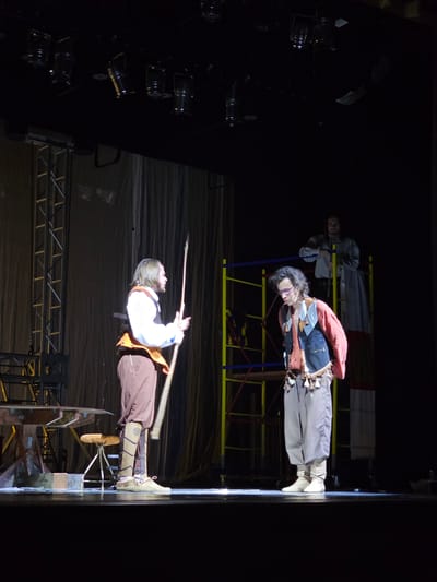 Two individuals, dressed in historical costumes, are the subjects of this image. The person in the foreground is a man with long, dark curly hair, wearing a red jacket over a dark vest and brown pants, with knee-high boots. He is holding a long pole or staff. The second person, in the background and slightly above, appears to be a woman in a light-colored dress or tunic. They are positioned on a stage with theatrical lighting equipment visible on the left side of the frame. The stage itself has a dark background and appears to be part of a theatrical production, possibly a play or performance. The overall mood is dramatic, with spotlights illuminating the performers. The time of day is likely evening or night due to the stage lighting and dark surroundings. There is no visible text in the image.