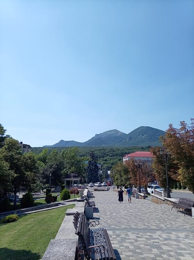 The image shows a view of the city of Zheleznovodsk, Russia, looking towards the mountains. In the foreground, there is a paved walkway lined with benches and manicured greenery. Several people are walking along the path, and a gazebo is visible in the distance. Beyond the park area, buildings and trees are seen, with a range of mountains forming the backdrop under a clear blue sky. The overall scene depicts a peaceful urban park setting with a natural landscape.
