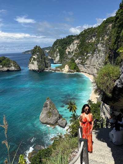 The image shows a vertical view of a scenic landscape in Pejukutan, Indonesia. Dominating the scene is a turquoise bay with rocky cliffs and verdant vegetation. A woman in an orange dress and sunglasses stands near a wooden railing along the edge of the cliff, posing for the camera. Palm trees are near the edge of the bay. The sky is blue with some clouds. In the distance, rock formations jut out of the sea. The turquoise water contrasts with the gray rock formations and the sandy beach.