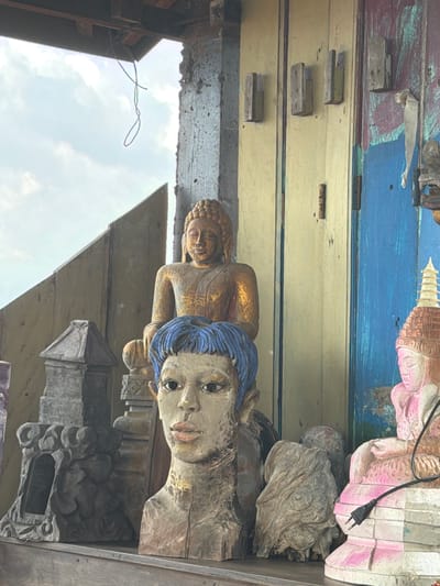 The image shows a close-up of various wooden sculptures displayed outdoors, likely on a porch or shelf, in front of a weathered wooden structure. The sculptures include a golden Buddha statue, a bust of a person with blue hair, and another carved figure in pink and white. In the background, a blue and yellow wooden door or wall is visible. There is a black electrical plug resting on the pink and white sculpture. The sky is visible in the upper left corner, with clouds. The setting appears to be an outdoor artisan stall or shop, possibly in Tejakula, Indonesia, given the style of the carvings and the weathered materials. The lighting suggests it is daytime, with natural light illuminating the scene.