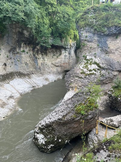 This is an aerial, high-angle shot of the Khadzhokh gorge in Kamennomostskiy, Russia. The gorge is characterized by steep, rocky cliffs covered in lush green foliage. A muddy, fast-flowing river cuts through the bottom of the gorge. Large boulders, some covered in moss, are scattered throughout the scene. In the upper right corner, a small group of people can be seen standing on the edge of the cliff, observing the gorge. A yellow railing is visible on the right side of the image, providing a safety barrier along the cliff edge.