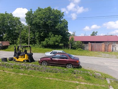 In Atabe, Georgia, under a bright, cloudy sky suggesting midday, a rural scene unfolds. In the foreground, a vibrant green lawn slopes upwards, bordered by a low, dirt embankment. To the left, a stack of old tires lies on the grass next to a yellow forklift, which is partially covered by a black tarp. The forklift bears Cyrillic text, "ДП-1504". Beyond the forklift, a dark red sedan is parked, its wheels turned slightly. Further back, a lighter colored sedan is parked behind it.
The middle ground features a gravel road that curves towards a large, red-roofed building with wooden double doors and brick accents. The building appears to be a barn or workshop. Behind the building, lush green trees dominate the landscape, their dense foliage reaching towards the sky. Power lines crisscross the upper portion of the image, a common sight in rural and semi-urban settings. The overall impression is one of a quiet, slightly overgrown rural area, possibly a farm or industrial yard on a sunny day. No people are visible in the image.