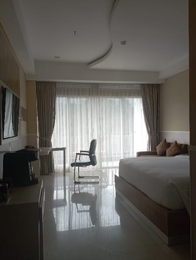 The image captures an interior room, identified as being in Batulawang, Indonesia. The room features a large window or glass door at the far end, covered by sheer white curtains and beige drapes, allowing bright natural light to flood the space and reflect off the glossy, light-colored tiled floor. On the left, a dark credenza or cabinet supports a black coffee machine and additional dark objects, with a wall-mounted television partially visible above it. Adjacent to this, a small, light-toned desk is positioned with a dark office chair. To the right, a bed with white linens and a wooden frame is prominently situated. A section of a brown sofa with two light brown throw pillows is visible behind the bed on the right. The white ceiling displays a decorative curved element, recessed light fixtures, and a smoke detector. No individuals are present.