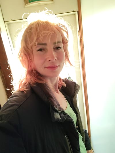 A medium shot of a woman with pink hair and makeup is captured from the chest up. She is wearing a black jacket over a light green t-shirt. The background is blurred, with bright light emanating from the right side of the frame, suggesting an outdoor setting or a room with a large window or light source. The overall lighting of the image is warm and bright, with the sunlight backlighting the woman's hair, creating a halo effect. The woman is looking at the camera and smiling slightly. The image quality is average, with some graininess and minor focus issues. The location in Shcheleiki, Russia is not discernible from the visual information provided in the image.