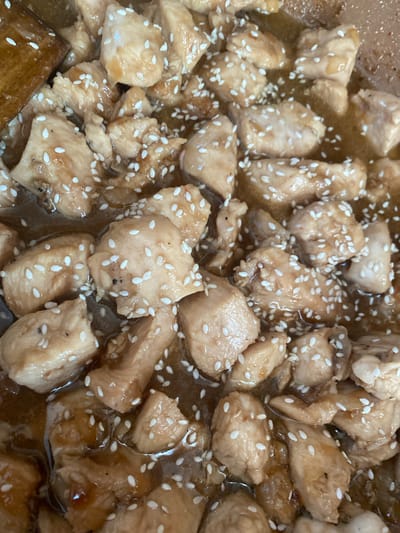 The image is a close-up, overhead view of bite-sized pieces of cooked chicken coated in a glossy sauce and sprinkled with white sesame seeds. The chicken pieces vary in shape and size, appearing to be lightly browned and tender. The sauce is a rich, brown color, reflecting the light and coating the chicken evenly. White sesame seeds are scattered liberally across the dish, adding a textural contrast. The background is blurred, but suggests a cooking environment, possibly a pan or wok, with a hint of a wooden utensil visible on the left edge of the frame. The image is brightly lit, highlighting the glossy texture of the sauce and the individual sesame seeds. There is no indication of a specific location in Kisliakovskaya, Russia.