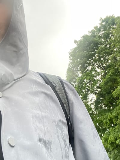 This is a first-person perspective shot taken outdoors in Bol'shoe Zaborod'e, Russia. The shot is tilted, with the upper portion dominated by the green foliage of a tree against a bright, overcast sky. The foreground is occupied by a person wearing a light gray, hooded jacket. The jacket appears to be made of a waterproof material, as evidenced by the water droplets and streaks visible on its surface, suggesting it is raining or has recently rained.
A black and gray textured band with the "VANS" logo is visible around the person's waist or chest area, likely part of a backpack or hydration pack. The lower part of the image shows the collar of the jacket and two light-colored circular buttons. The weather appears to be dull and possibly misty, given the bright, diffused light and the wetness on the jacket. The overall impression is that the person is outdoors, possibly hiking or walking, and caught in inclement weather. The scene is natural and appears to be a casual moment during outdoor activity.