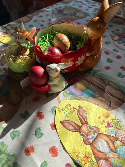 The image shows an Easter-themed still life on a patterned tablecloth. The tablecloth has a white base with a repeating pattern of orange flowers and green leaves. There are also larger images of brown rabbits and Easter eggs on the cloth.

Several Easter decorations are arranged on the tablecloth. There is a ceramic brown bunny figurine resting on the edge of a red ceramic dish with white daisy accents. Inside the dish are colored Easter eggs and green shredded paper. Next to the red dish is a white ceramic bunny figurine lying next to a red Easter egg. Below these items is a green and yellow Easter egg-shaped container holding a smaller bunny figurine.

A yellow napkin with a rabbit image and the words "Happy Easter" is placed to the upper right of the frame.

There are no people visible in the image. The location is not discernible from the image, and there is no evidence to suggest it is in Tompsan, Bulgaria.