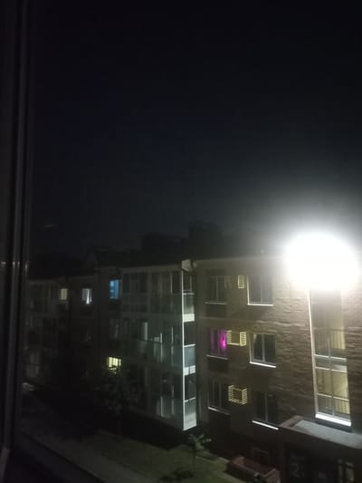 This is a nighttime, eye-level, exterior shot of an apartment building, likely in the city of Novoye Shigaleevo, Russia, as indicated by its architectural style and the presence of a streetlamp. The building is constructed of brick and has multiple floors with numerous windows. Some windows are illuminated, indicating occupancy. A bright, glaring streetlamp is positioned to the right of the frame, casting a strong light on the building and creating a lens flare. The sky is dark, suggesting it is late at night. In the foreground, on the left, is a vertical element, possibly a window frame or part of the building's exterior. The ground in front of the building appears to be paved, with some trees and greenery visible. There are no people or vehicles clearly visible in the frame.