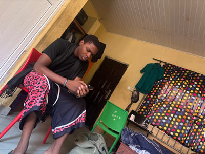 The image depicts an indoor scene within a room in Ado-Ekiti, Nigeria. A male individual, appearing young adult, is seated on a red plastic chair, looking down intently at a dark-colored smartphone held in both hands. He is wearing a black short-sleeved t-shirt, a black scarf draped over his left shoulder and lap, and dark shorts that feature a red and black leopard print pattern on the upper part, transitioning to solid black with white stripes on the lower part. A dark-colored bracelet is visible on his left wrist. He is barefoot, with a dark flip-flop partially visible on the floor below his left foot.

To the right of the seated individual, a green plastic chair is positioned, bearing white markings "F C215 8" on its backrest. The walls of the room are painted a light yellow-beige, showing some signs of wear. A dark-colored wooden door is visible on the left side of the frame. On the right wall, a dark green short-sleeved collared shirt hangs from a black rod or hanger, with a dark cap suspended beneath it. Adjacent to this, a multi-colored polka-dot curtain with a black background and dots in yellow, orange, red, blue, and green, covers an opening. Below the curtain, a dark-colored shelf or table holds a white cup and other indistinct items. A bed with blue and white patterned bedding is partially visible in the lower right, and a light-colored fabric bag rests on the floor in front of it. The ceiling is white and constructed from horizontal slats.