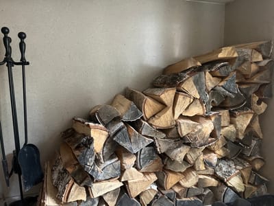 A pile of firewood sits against a light-colored wall in an indoor space. To the left of the firewood, there is a black set of fireplace tools, including a poker, tongs, and shovel. The firewood appears to be birch logs. The location of the image is not discernible as Patrony, Russia, from the visual information.