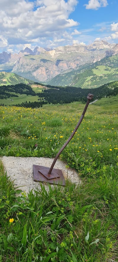 An abstract sculpture, resembling a bolt with two nuts, stands in a grassy, mountainous landscape. The sculpture is made of rusted metal, with a square base and a long, curved rod topped with two hexagonal nuts. It is positioned on a concrete pad surrounded by lush green grass dotted with small yellow flowers. In the background, a vast expanse of rolling hills and rugged mountains unfolds under a partly cloudy blue sky. The mountains exhibit a rugged, rocky texture with patches of snow near their peaks, and are covered in dense evergreen forests on their lower slopes. Further in the distance, there are signs of human activity, including buildings and ski lifts, suggesting a resort area. The overall scene depicts a blend of natural beauty and human intervention in a high-altitude environment, likely in the Dolomites region of Italy, near Sëlva - Wolkenstein - Selva di Val Gardena. There are no people visible in the image.