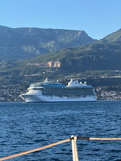An overcast, clear day captures a large, white cruise ship named "Allura" sailing on calm blue water. The ship is positioned in the middle ground of the image, with its bow facing left. The ship is multi-decked with many windows, and its upper decks are a light blue color. In the background, a densely populated coastal city with buildings of various sizes and colors is visible, nestled at the base of a rugged, green mountain range. The mountains are steep and rocky, with some bare patches of earth, suggesting quarrying or landslides. The sky is a clear, pale blue. In the foreground, a portion of a boat, with ropes and railing, is visible, indicating the photograph was taken from another vessel.