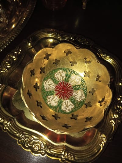 A small, ornate golden bowl sits atop a larger, square, golden tray. The bowl features a scalloped edge with cross-shaped cutouts, and a floral design in the center with red, white, and green details. The tray, also golden, has an elaborate, raised rim. The lighting is dim, casting shadows across the items. A blurred golden dish is partially visible in the upper left corner.