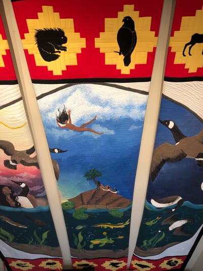This image is a photograph of a ceiling quilt at the Orillia Public Library in Severn, Canada. The quilt is divided into panels, each depicting a scene with animals and a human figure. The top panels feature silhouetted animals against a red and yellow geometric background, including a porcupine, a bird, and a deer. The lower panels depict more detailed scenes. One panel shows a woman falling from the sky into a blue expanse, surrounded by geese and aquatic life. Another panel features a large turtle supporting a small island with a tree and a reclining human figure, with geese flying overhead. The bottom of the quilt has a border with repeating animal motifs. The quilt is illuminated by natural light, casting shadows from the visible beams supporting the ceiling.