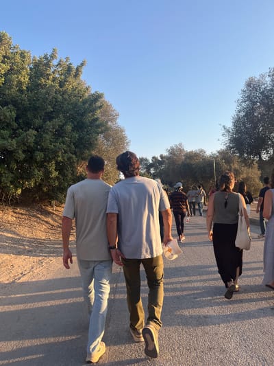 The image depicts an outdoor scene with multiple individuals walking away from the camera along a paved path under a clear blue sky. The setting is identified as the Brener Regional Council, Israel.
In the immediate foreground, two men walk side-by-side. The man on the left wears a light beige short-sleeved t-shirt, light blue jeans, and light athletic shoes. The man on the right wears a light grey short-sleeved t-shirt, olive green pants, and light-colored athletic shoes. He has dark, wavy hair and holds a white baseball cap with an orange/yellow logo in his left hand. A dark wristband or watch is visible on his left wrist. Their proximity suggests they are walking together.
Further along the path on the right, a woman is visible from behind, wearing a dark green sleeveless top with a cut-out detail on the back, a long dark skirt, and carrying a light beige tote bag on her left shoulder. Numerous other individuals and small groups are visible further down the path, all walking in the same direction. One person wears a dark and light striped shirt and a white cap. A plastic bottle with a pink cap is discernible on the ground near this group.
The path is light-colored asphalt, with a lighter dirt or gravel shoulder on the left side. Lush green trees and bushes with dense foliage line both sides of the path, creating a verdant environment. Long shadows of the people extend towards the camera on the path, indicating the sun is low behind them, characteristic of late afternoon or early evening. A street light pole is partially visible in the distance among the trees on the right. The sky is uniformly clear and bright blue, devoid of clouds.