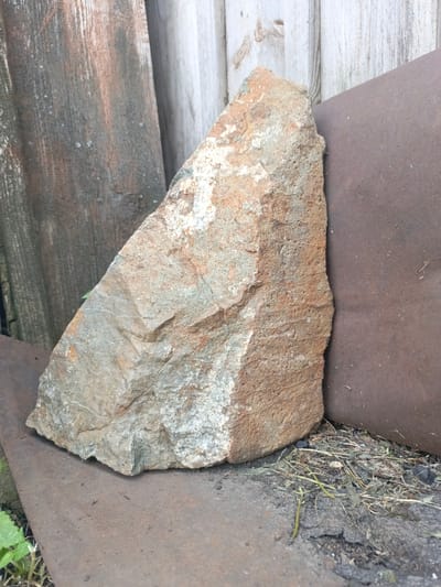 A close-up, outdoor, eye-level shot shows a large, rough, triangular-shaped rock with varied earthy tones of brown, gray, and white. The rock is positioned leaning against a dark brown, curved metal surface on the right. To the left and behind the rock, weathered wooden planks with vertical grain patterns form a rustic backdrop. The ground beneath the rock and metal surface consists of dry grass, dirt, and cracked pavement. The overall scene evokes a sense of abandonment or disuse. There is no indication of the city of Zalesovo, Russia within the image.