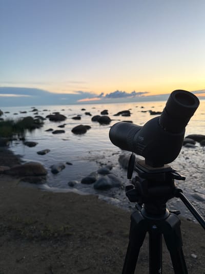A black spotting scope, mounted on a black tripod, is positioned on a sandy and rocky shore at the edge of a body of water. The spotting scope is angled towards the water and the horizon, where the sky transitions from a pale blue to a soft orange and yellow as the sun sets. Scattered rocks and some sparse vegetation are visible along the shoreline. The water is calm, with gentle ripples reflecting the colors of the sky. In the distance, more rocks emerge from the water, and a few wispy clouds are present on the horizon. The scene is serene and peaceful, likely captured in Haabneeme alevik, Estonia, given the characteristic coastal landscape.