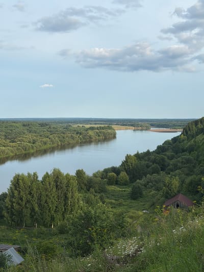 This image depicts a scenic view of a wide river flowing through a lush, green, forested landscape. The river curves gently, with its water reflecting the pale blue sky and scattered clouds. The foreground features a grassy hillside with wildflowers and some trees. In the mid-ground, a dense forest covers the rolling hills on either side of the river. On the right side of the frame, nestled within the trees, is a rustic, abandoned wooden building with a dark, reddish-brown roof. In the distance, the river opens up to a wider expanse, with more trees and a flat plain stretching to the horizon. The overall impression is one of natural beauty and tranquility, with a touch of rural abandonment. The location is the city of Cherdyn, Russia, specifically a view overlooking the Kama River.