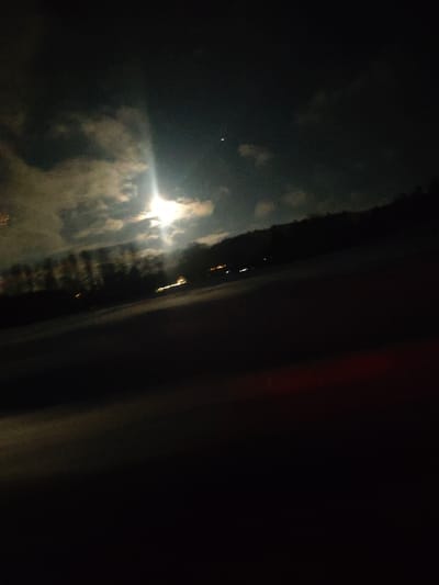 A nocturnal scene reveals a bright moon shining through scattered clouds, casting a luminous glow. Beneath the sky, a darkened landscape shows a row of trees and faint lights along the horizon. The lower portion of the image is predominantly dark, suggesting a foreground of shadow or undefined terrain. The slight tilt of the photograph adds a sense of movement.