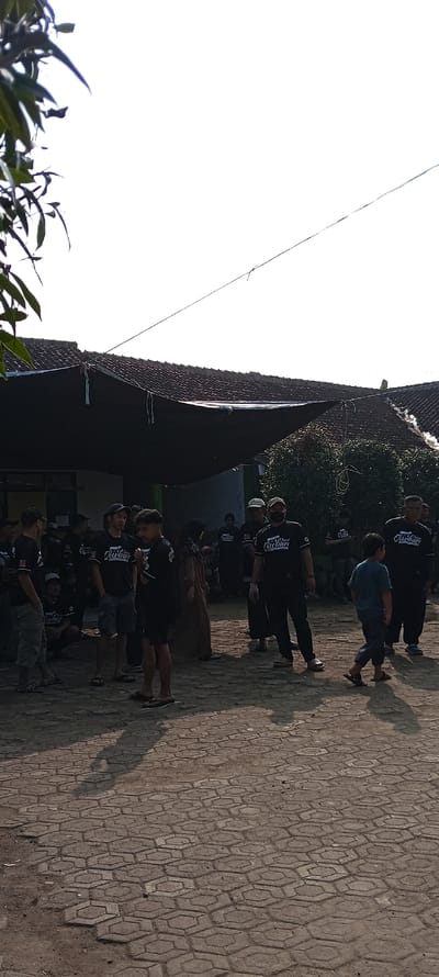 The image shows a group of people standing on a paved area in front of a building in Cangkuang, Indonesia (latitude: -7.065, longitude: 107.552). The building is partially obscured by a black tarp, which is stretched over the roof and supported by poles. There is a tree on the left side of the image, and a power line runs across the sky. The group of people appears to be gathered for some event or activity, as they are all wearing similar clothing and some are holding objects. The people are mostly men, with a few women and children present. The ground is covered in hexagonal patterned paving stones, with dirt and dust visible in the spaces between the stones.